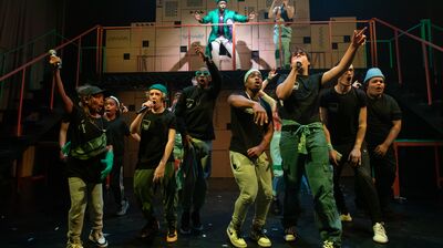 12 performers beatbox on stage with a raised balcony and cardboard cityscape. The performers wear black and green costumes.