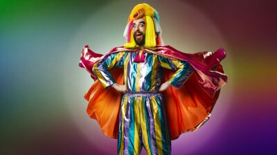 Person wearing a colourful striped jumpsuit with a cape flying out behind them. Their hands are on hips and they stand against a rainbow coloured background.