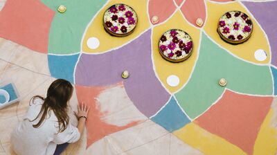 A person creating Rangoli artwork, with a multicoloured pattern with bowls of pink and white flowers placed on top