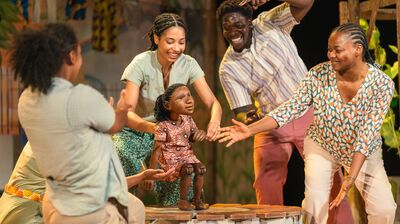 A group of performers wearing colourful costumes dance around a small puppet of a young girl.