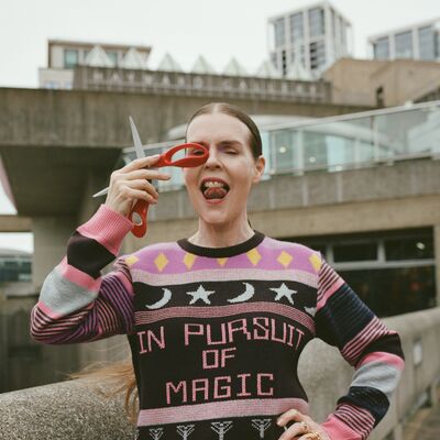 A white woman stands outside on a grey day; she is looking through the handle of a red pair of scissors wearing a knitted jumper saying 'In Pursuit of Magic'