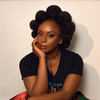 Chimamanda Ngozie Adichie resting her head in her hand, wearing a green and red satin skirt and black t shirt