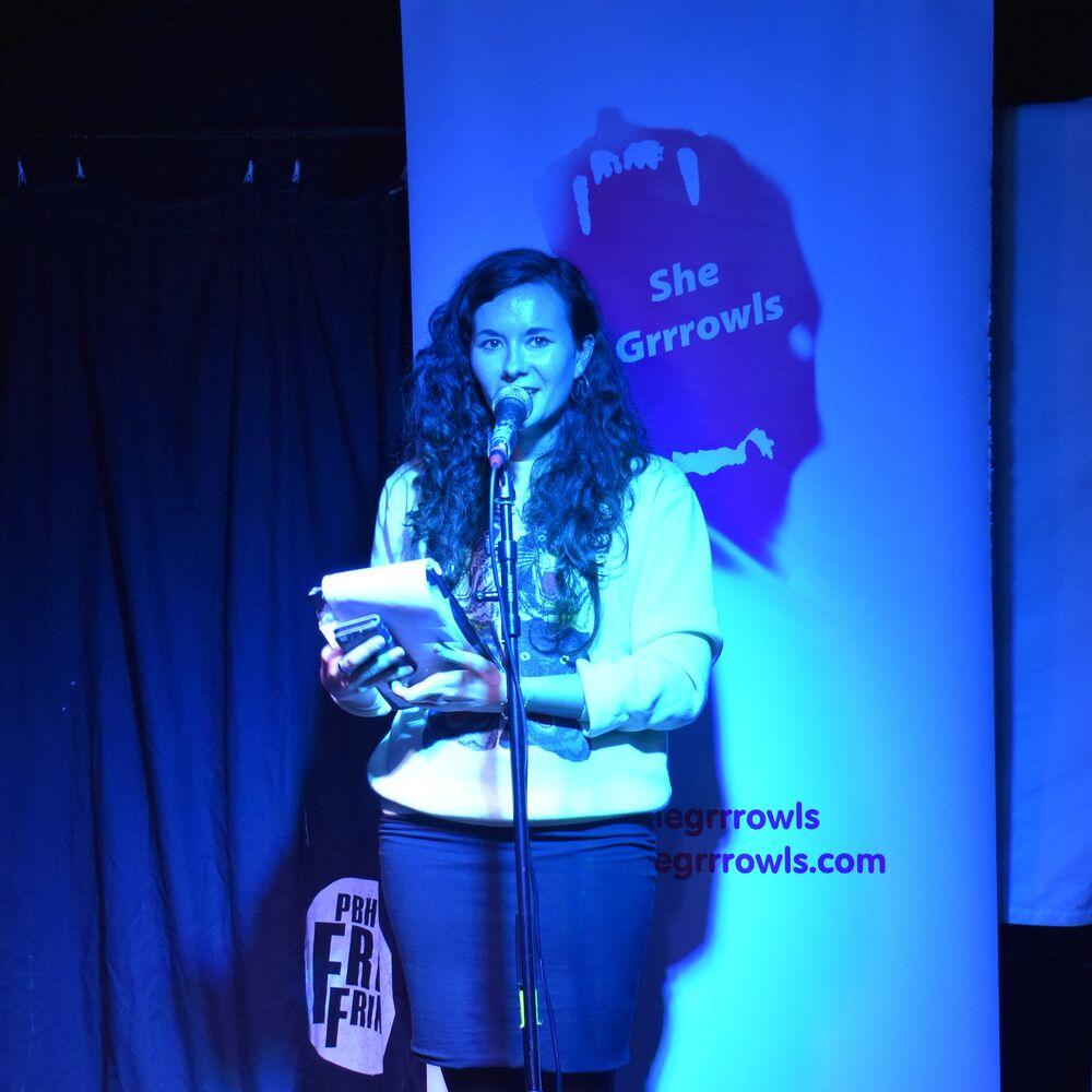 Woman stands at a microphone onstage against a blue background.