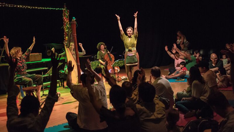 A live performance where musicians, including a harpist and a cellist, share the stage with a central performer in an intimate setting. The audience, seated on the floor, is actively participating by raising their hands, creating a sense of shared, joyful engagement with the artists.