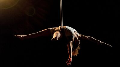A circus performer hangs in a dark space, suspended from a silk, their arms and legs fully extended
