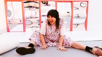 A smiling woman with dark hair and bangs wearing a pink floral dress sits on the floor with legs spread in front of several pink-framed funhouse mirrors.