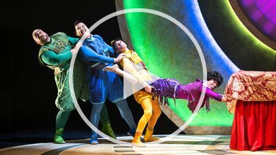 Four brightly costumed acrobats or actors, in a human chain, strain dramatically against a low red table and a backdrop of large, colorful glowing arches on a stage.