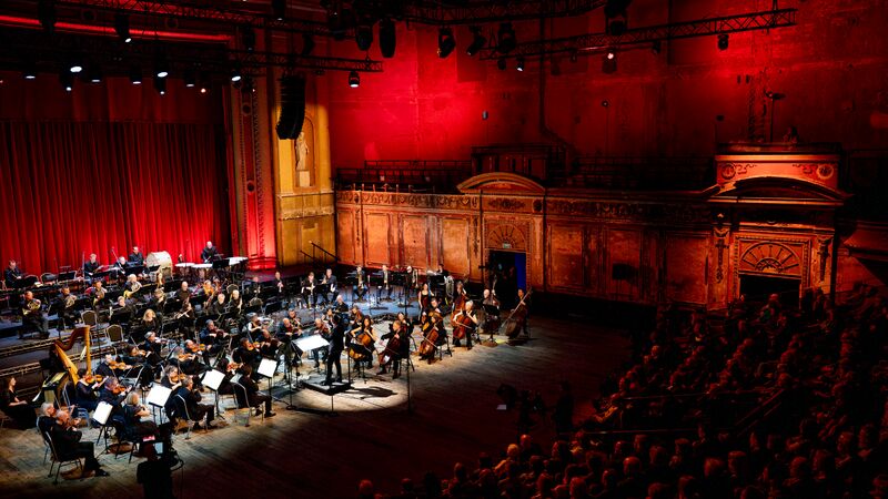 Concert Orchestra on stage under stage lighting in a large auditorium with wooden floors