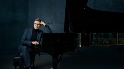 A person in a navy suit, black turtleneck and blue glasses sat and leaning against a grand piano