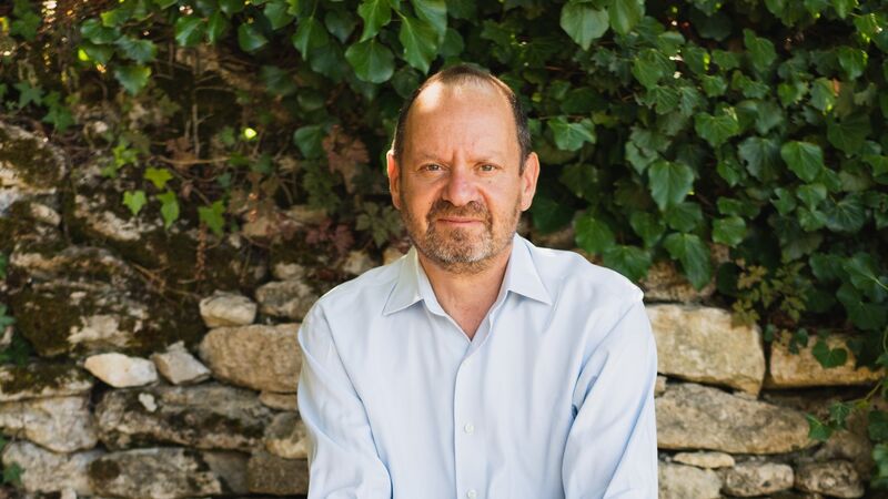 Philippe Sands sitting on a bench against a leafy background.