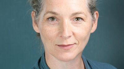 Ita O'Brien wearing a blue top against a teal background.