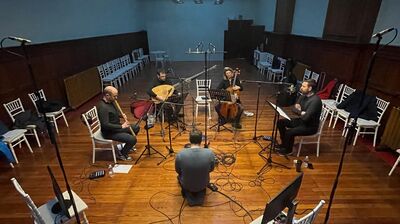 four people sitting on chairs with instruments in open recording studio with one man taking a video