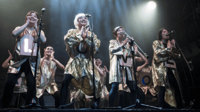 Four singers stood on stage with microphones weaing gold outfits.