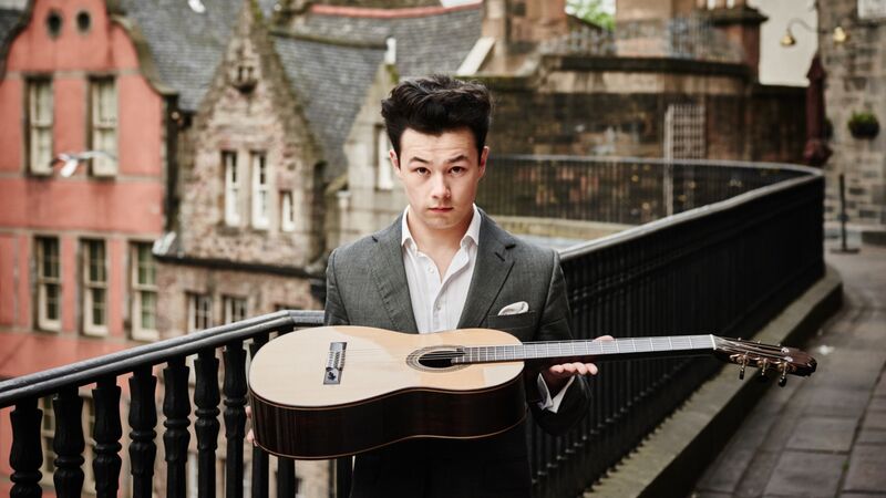 A man holding a guitar, standing on a balcony