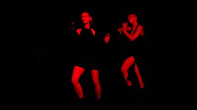 Two people wearing black on a black stage lit with red stage lighting moving and dancing