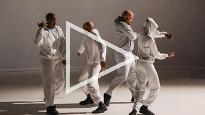 Four dancers wearing all white tracksuit outfits in a large white room they are all moving