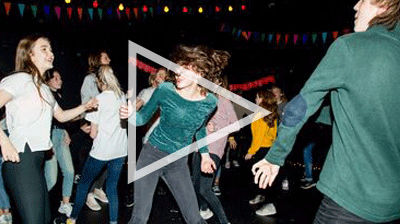 Group of young people dancing in a dark room with bunting and fairy lights, they are smiling and talking