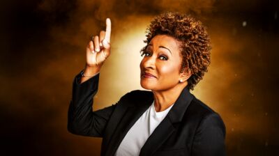 Wanda Sykes wearing a white t shirt and black blazer, pointing up and smiling