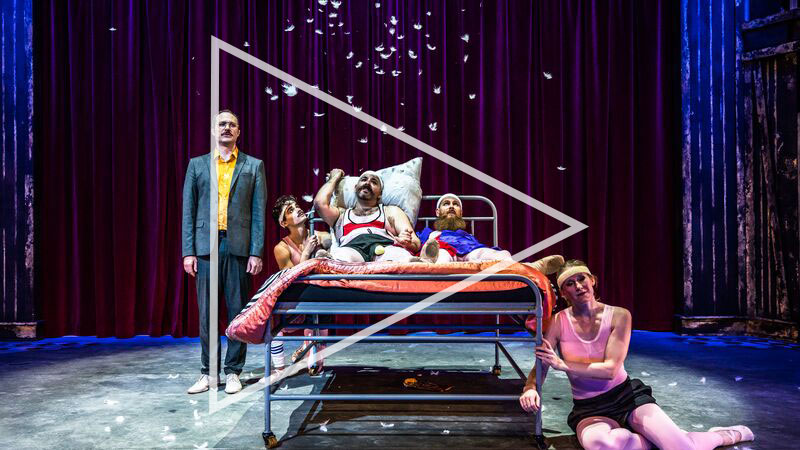 Six performers wearing pink leotards, black tutus and masks, stand in a line and perform ballet choreography.