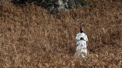 Ghetts is wearing a white patterned jacket and is standing in an area of brown dead ferns with rock behind him