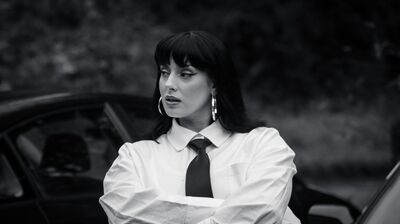 A person with dark hair and fringe wearing a shirt and tie with their arms folded, leaning against a car