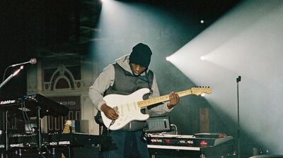 A man plays a white guitar surrounded by keyboards and other instruments