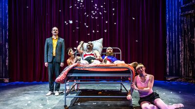 two performers lie in bed while other performers gather around them. White feathers fall onto the bed.