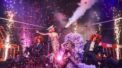 Performers on stage surrounded by falling confetti and they are wearing gold dresses or suits and large red wigs