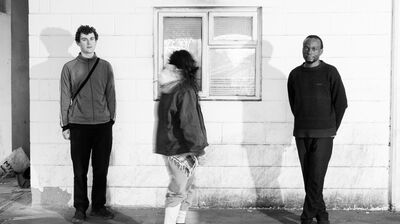 Three people standing outside a white wall, two looking at the camera one facing away