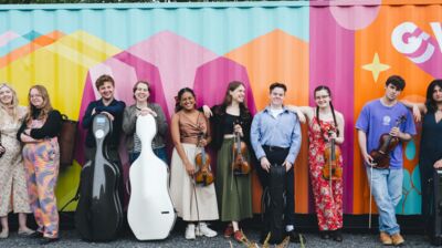 Orchestra musicians standing in front of a colourful shipping container holding a variety of instruments