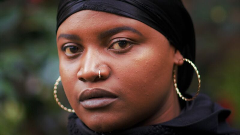 A person wearing black and large gold hoop earrings and a nose ring. They have deep brown eyes