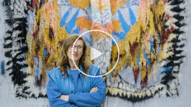 Artist Caroline Achaintre, a middle-aged woman with long wavy brown hair, wearing glasses and a powder blue sweatshirt and sleeveless jacket, stands in front of one of her multi-coloured woven artworks