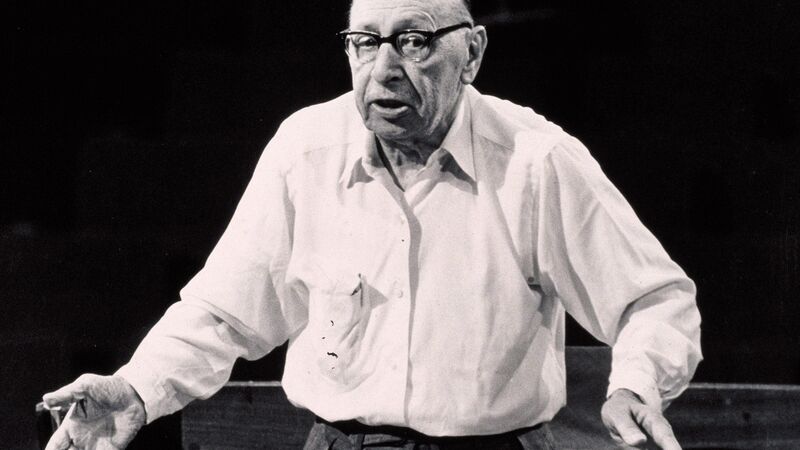 Igor Stravinsky, a small 83 year old balding man wearing black-rimmed glasses stands with arms stretched out at his stands as he conducts a rehearsal