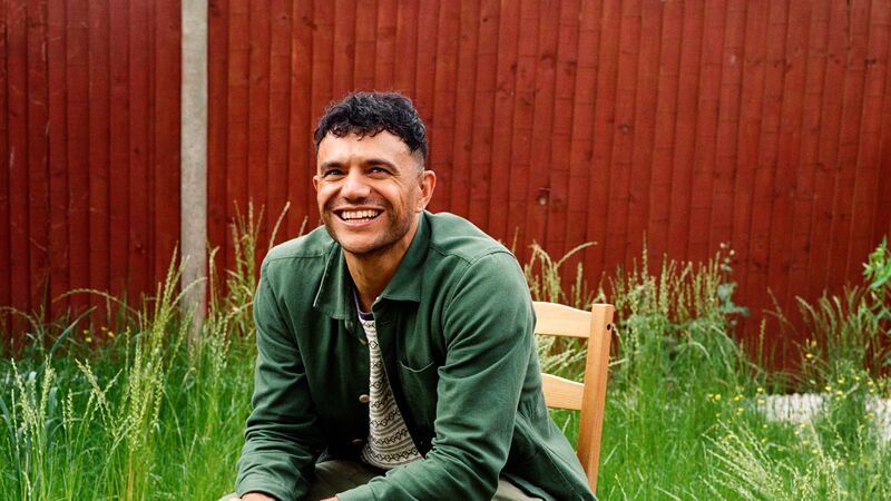 The poet Raymond Antrobus, a man in his late 30s of British and Jamaican heritage with short hair, sits smiling on a wooden chair in a roughly kept garden