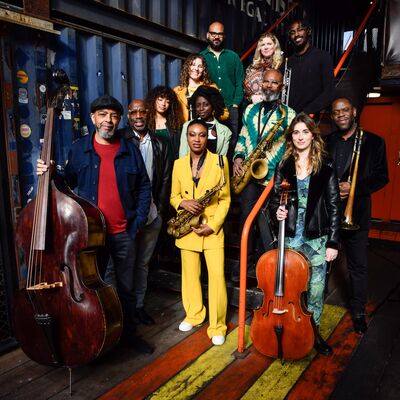 Jazz Jamaica, a band of 12 musicians stand in an industrial backdrop with their instruments