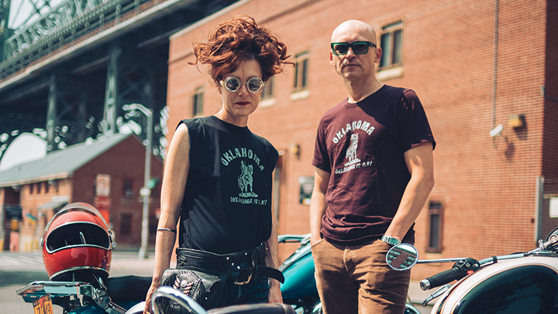 Theatre directors Kelly Copper – a woman with long auburn hair piled high on top of her haid – and Pavol Liska – a bald man wearing sunglasses – stand in between a couple of motorbikes wearing biker-style t-shirts; behind them is a brick building beside a metal suspension bridge