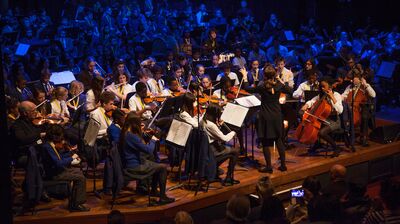 Group of secondary school musicians on a wooden stage seated in an orchestra layout with a conductor stood in the middle