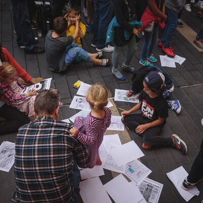 A group of children and adults draw on paper on the floor.