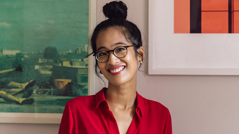 A woman in a red blouse sat smiling at the camera, she wears dark round glasses