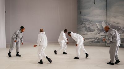 Five performers wearing grey and white tracksuits and black shoes moving with hunched over bodies