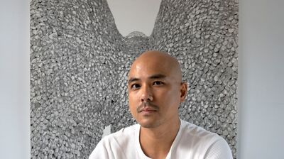 A person wearing white, looking to the camera with a silver artwork behind them and a drawing in front