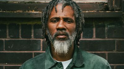 A man with a grey and white beard, wearing a green shirt looking to camera