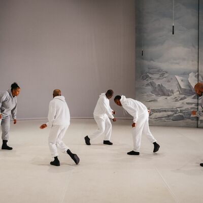 Five performers wearing grey and white tracksuits and black shoes moving with hunched over bodies
