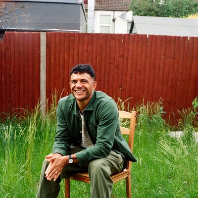 The poet Raymond Antrobus, a man in his late 30s of British and Jamaican heritage with short hair, sits smiling on a wooden chair in a roughly kept garden