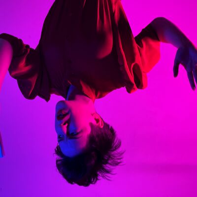 A an upside down person with hair hanging off their head gestures with their hands in front of a purple hued background.