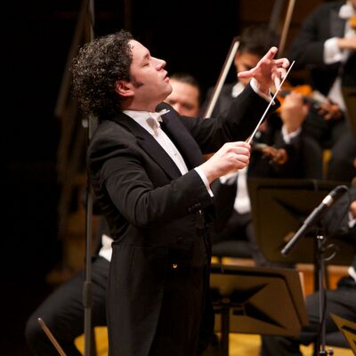 Conductor wearing a black suit white shirt and pink bow tie, with violinists playing around them