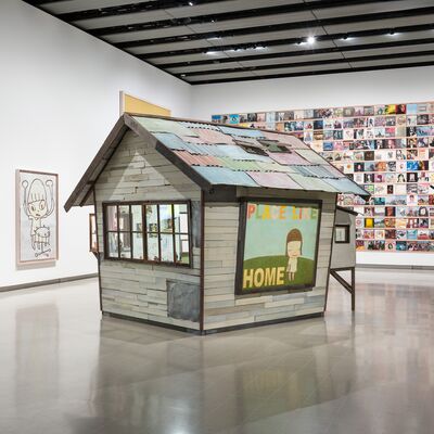 Sculpture installation of a small house with an artwork of a small figure next to the word home on the side in a large room with other artwork on the walls