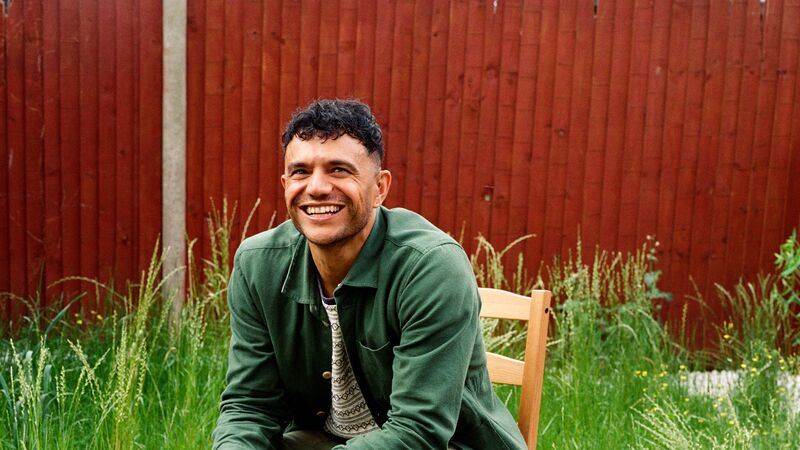 The poet Raymond Antrobus, a man in his late 30s of British and Jamaican heritage with short hair, sits smiling on a wooden chair in a roughly kept garden