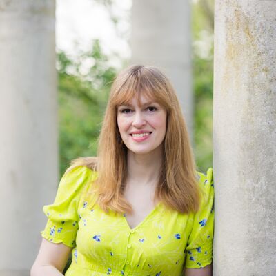 Elodie Harper wearing a yellow dress standing against a concrete column.