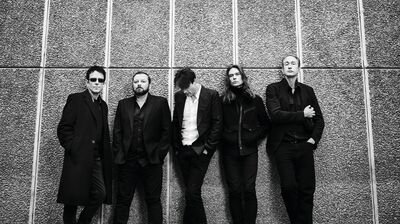 5 people wearing black smart outfits leaning against a concrete wall